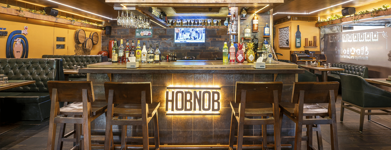 A bar counter with stools, a glowing 'HOBNOB' sign and bottles displayed behind at Infiniti Hotel, Indore.