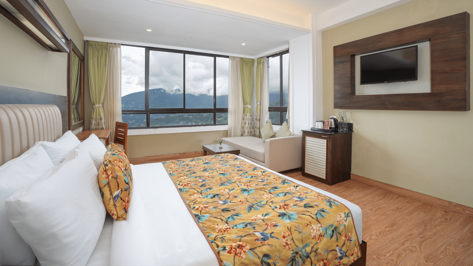Deluxe Valley View Room at Summit North Star, Gangtok featuring a bed, a flat TV screen, a cabinet table with tea and coffee maker, a sofa, a workstation and a large window.
