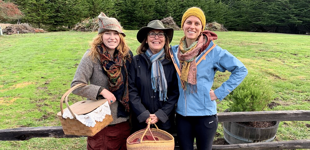 Three people standing together outdoors, holding baskets and smiling, with a fence and grassy field behind them.