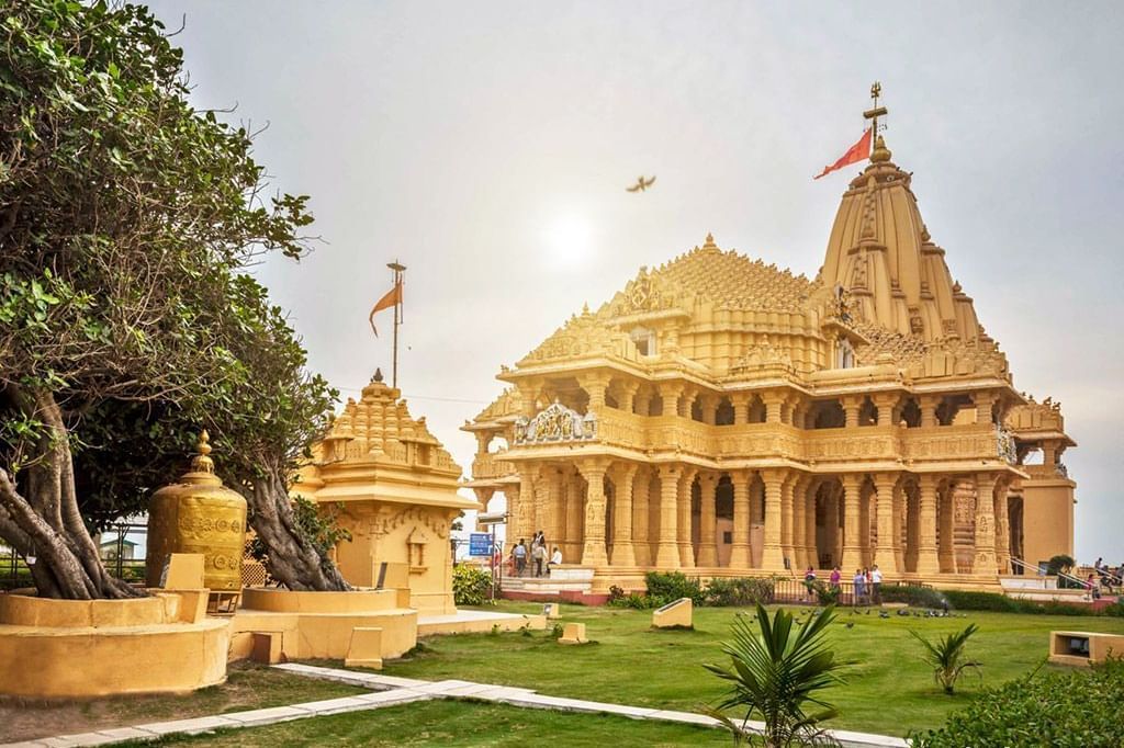 A grand sandstone Somnath temple with ornate carvings and fluttering saffron flags, surrounded by lush greenery.