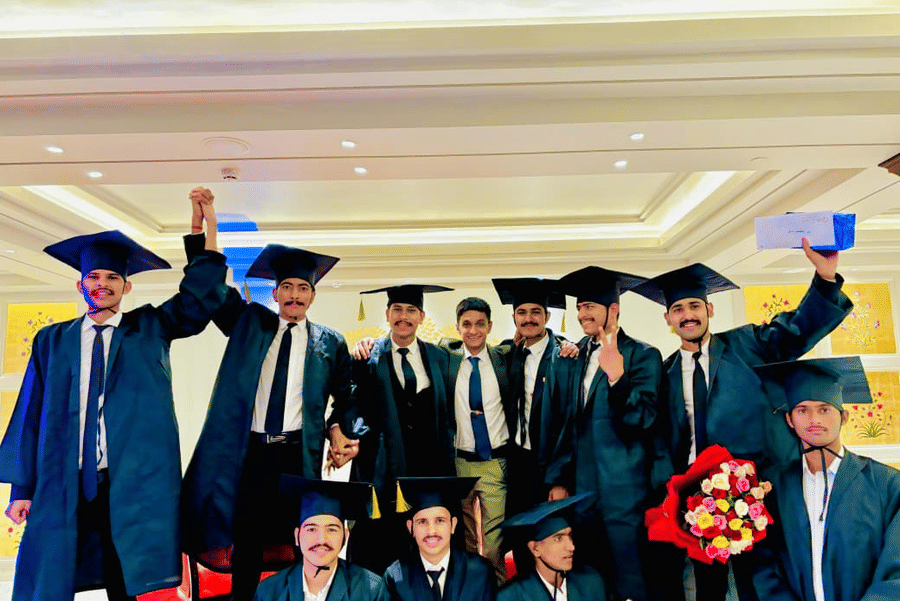 A group of graduates stands together indoors at Clarks Group of Hotels. They wear graduation gowns and caps while holding a bouquet and a box during the celebration.