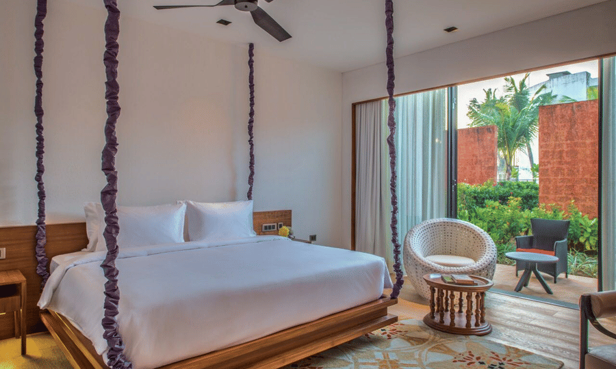 The Deluxe Patio King Room with a large wooden bed hanging from the ceiling on purple rope vines, and an armchair at Azaya Beach Resort, Goa.