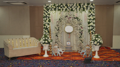 A decorated stage setup at Ambar Sarovar Portico, Gandhidham displays a patterned couch, floral arrangements, elephant figurines, and carpets.