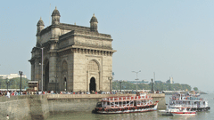 A stone gateway with boats in the harbour in front and people walking along the promenade.