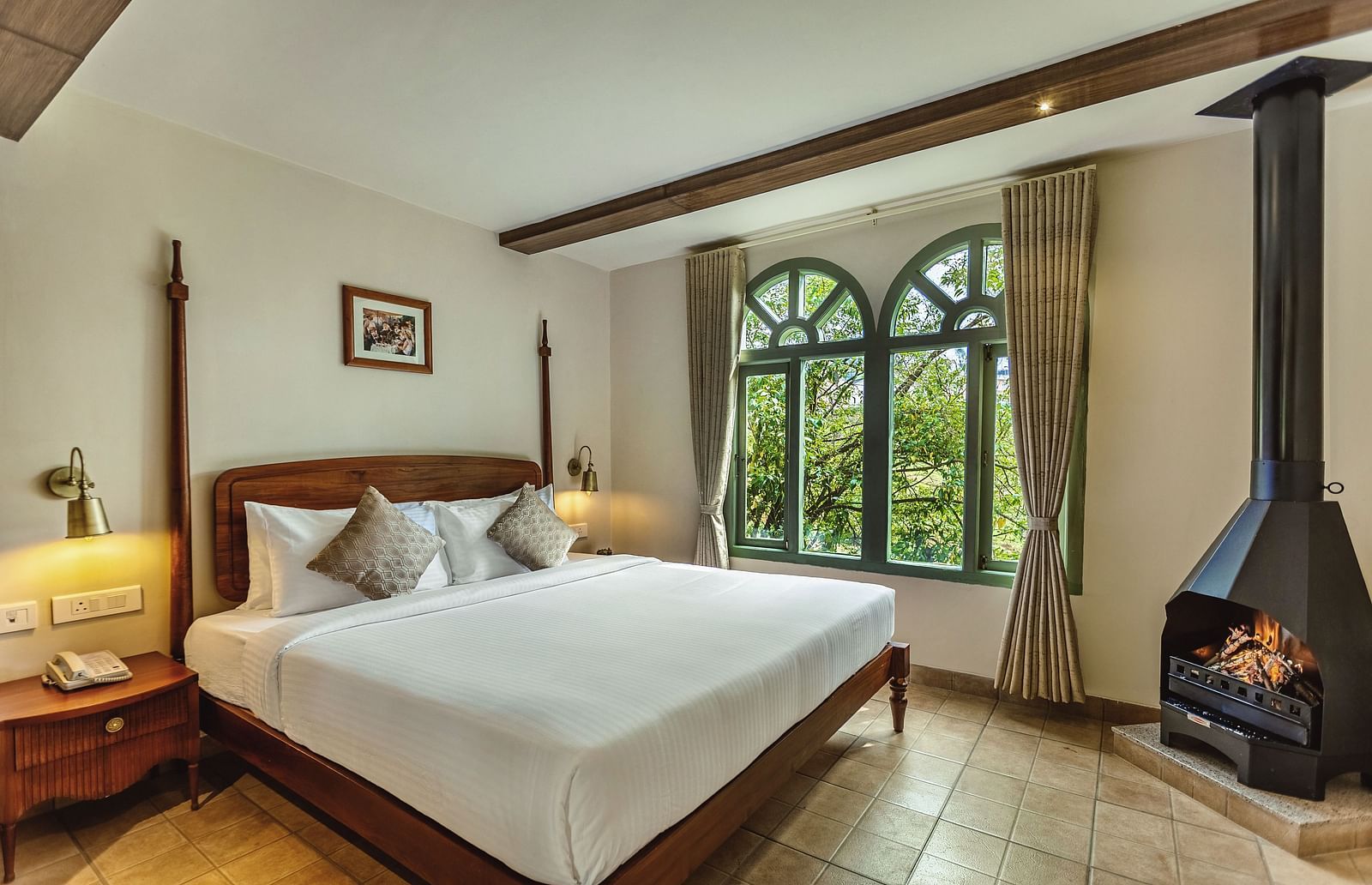 Sunlit bedroom at Mango Hill Shola, Ooty with a wooden bed, side tables, and large arched windows offering lush green views.