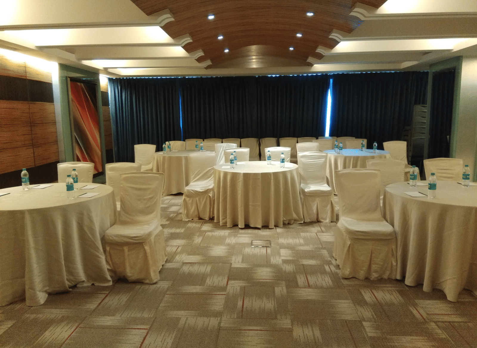 Banquet hall at Mirage Hotel, Mumbai featuring ample seating area, round tables covered in white silk covers and carpet flooring.