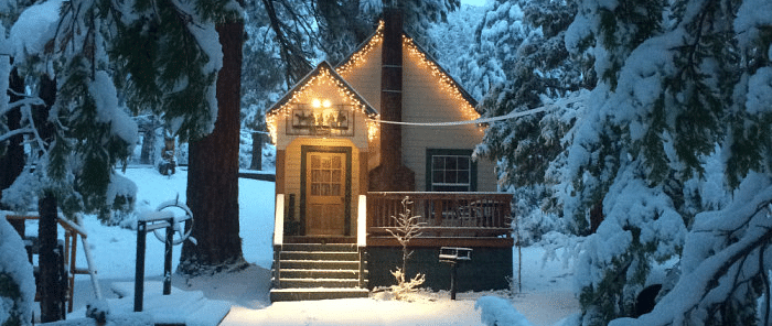 Snow covered cabin at Shaver Lake Village Hotel, with a lit doorway and porch, nestled among snow-laden pine trees.
