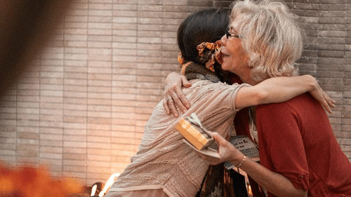 An older white woman hugs a younger woman who holds a small wrapped gift