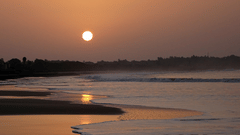 A sunset view over the sea with waves at the shore and mountains in the background.