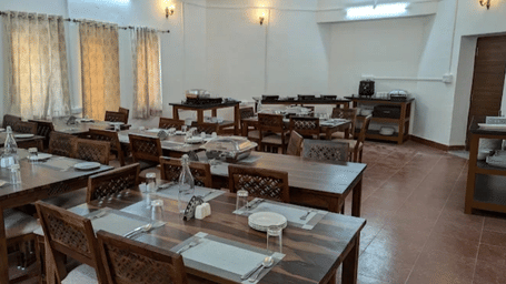 The dining area at Estherea Bagh, Ranthambore, is furnished with dark wood tables, chairs, and closed curtains.