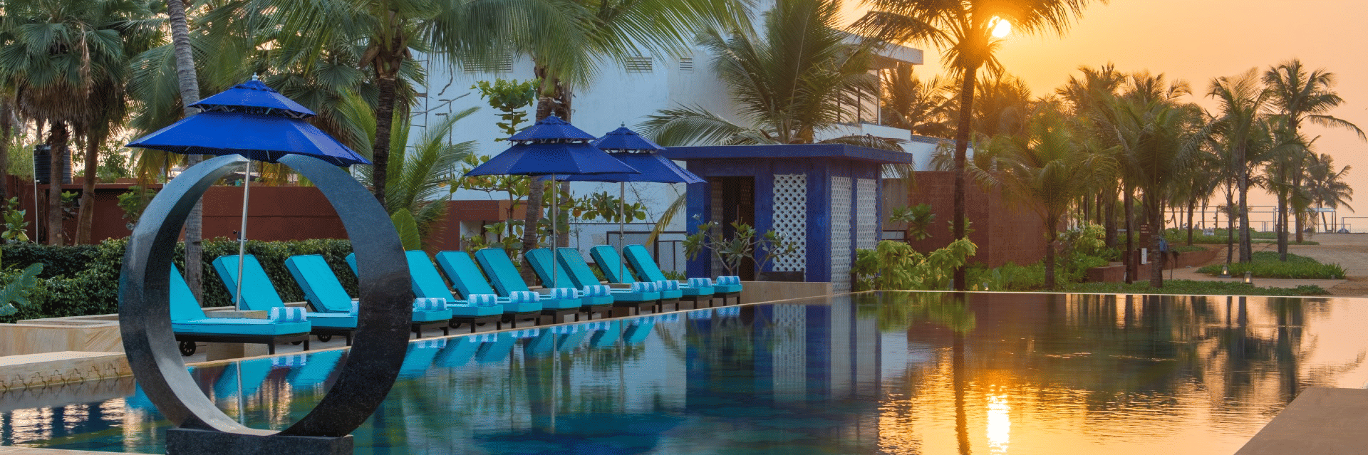 Swimming pool at Azaya Beach Resort in Goa and near to it there are lounger beds with the entire area surrounded by palm trees.