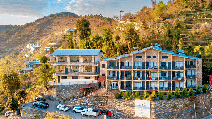 An aerial view of the multi-storey resort building situated on a green hillside under a blue sky at Himalayas Resort By The Lake Hill, Mukteshwar.