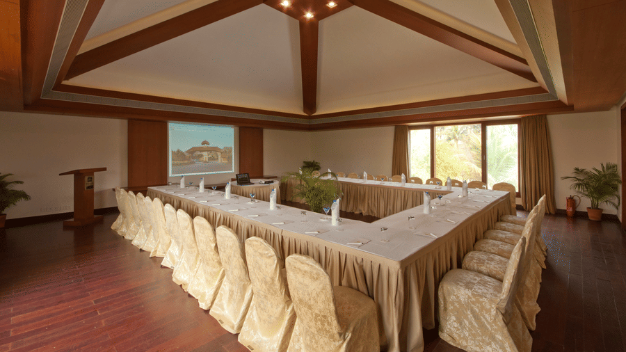 A conference room at Vedic Village Spa Resort featuring a U-shaped table setup with gold chair covers a high thatched ceiling wooden flooring and a projection screen at the front.