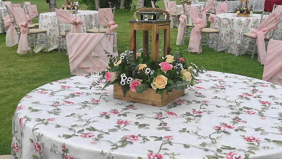 Round outdoor dining table with a floral tablecloth, topped with a wooden lantern and flower arrangements, set on a green lawn with other tables in the background - Black Thunder, Coimbatore