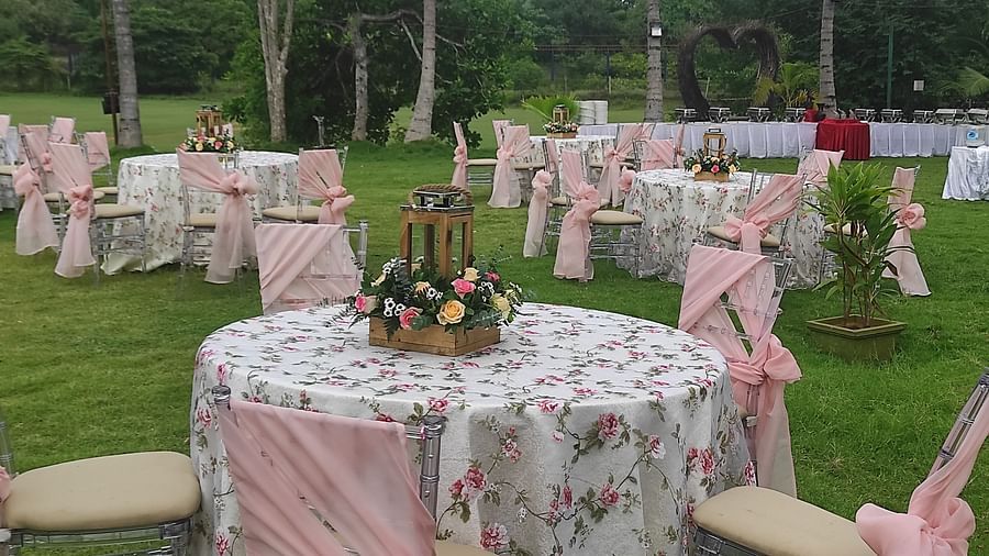 Outdoor dining setup on a lawn with round tables covered in floral tablecloths, chairs decorated with pink sashes, and a flower arrangement centrepiece - Black Thunder, Coimbatore