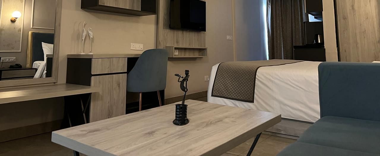 A spacious room featuring a large bed, a wooden table, and a study table with a chair in front of the wall, along with a wall-mounted TV at Urban Suites by BluSalzz, Jaipur.