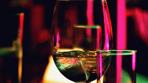 Wine glass and tumblers on a bar, illuminated by vibrant lights, creating a lively atmosphere.
