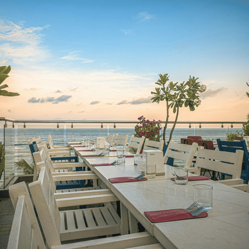 Open-air oceanfront restaurant terrace with white wooden tables and chairs set for dinner overlooking the sea at sunset