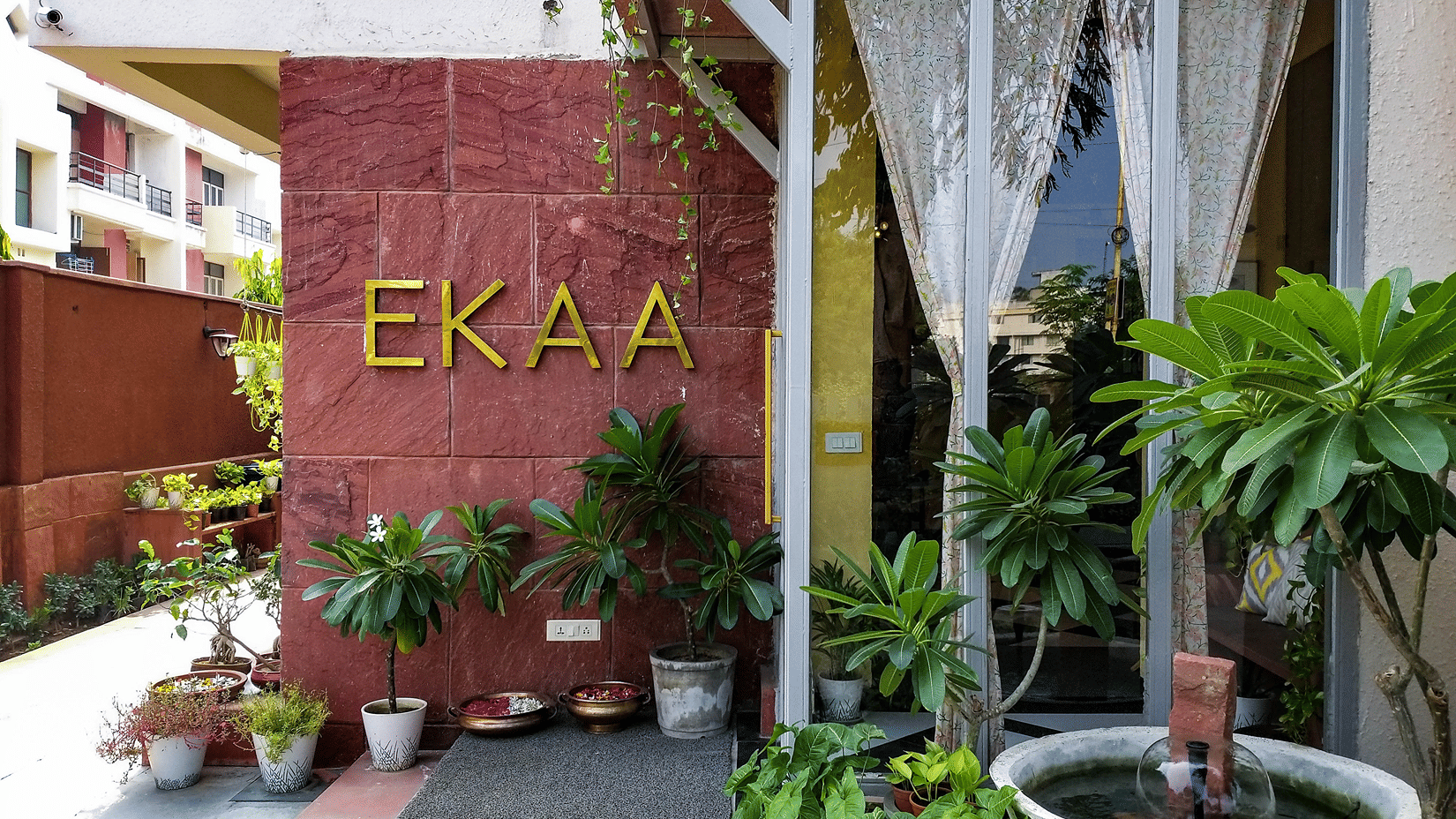 Exterior entrance view of EKAA spa featuring reddish stone walling with gold lettering, potted plants, and a small water basin near glass doors with white curtains.