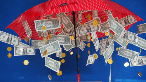 A close up of many notes and coins falling down from inside an open red umbrealla.