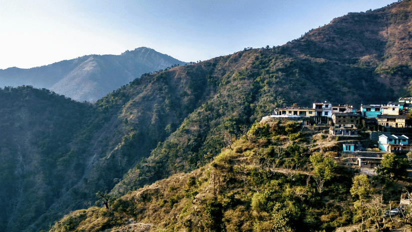 Houses on a mountain slope, surrounded by green ridges and distant hills under bright daylight in Dehradun, one of the best summer destinations in India