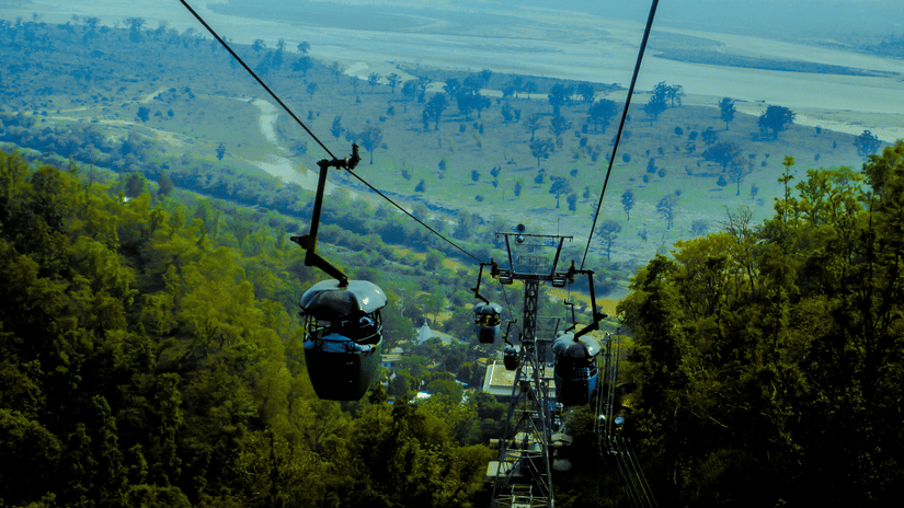 Ropeway moving over forested hills and valley, with distant plains visible under a hazy blue sky and mountainous backdrop