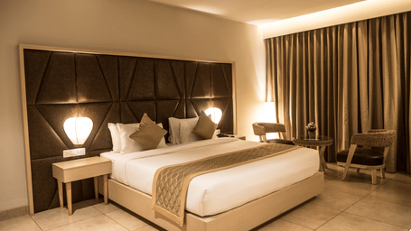 A Premium Room at Cholaa Dynasty, Kumbakonam, featuring a king size bed, matching nightstands and a seating area with 2 chairs and a table.