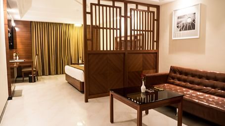 Interior view of a Premium Suite featuring a corner layout. A divider separates the living area with a sofa and tea table from the bedroom area, which includes a bed and a flat-screen TV at MM Legacy.