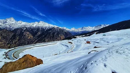 A snowy mountain landscape under a bright blue sky with wispy clouds, featuring a winding road and distant peaks.