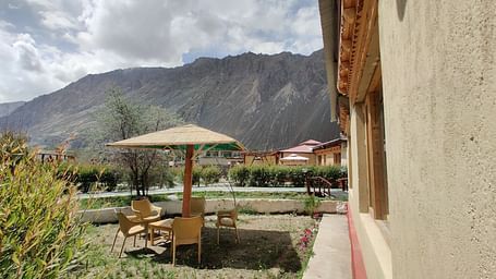 An outdoor seating area with a gazebo and chairs, adjacent to a building with a light-coloured exterior, with mountains visible in the background at White Sands Resort, Ladakh.
