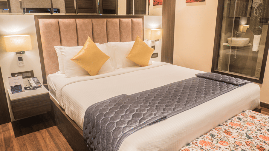 A Superior Room at Hotel Eastern Gate, Port Blair, featuring a neatly made bed, a nightstand, 2 wall lamps, and wooden flooring. 