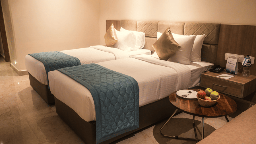 A Deluxe Twin Room at Misty Ocean, Pondicherry, featuring twin beds, a nightstand, and a basket of fruits atop a small table.