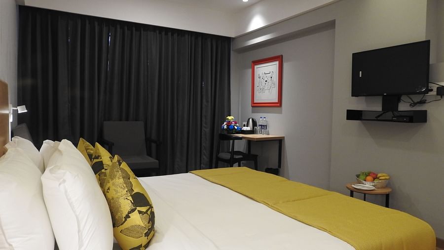 A hotel room featuring a double bed with gold pillows, a black armchair and a workspace with a television and fruit bowl - TGI Ameya Grand, Goa