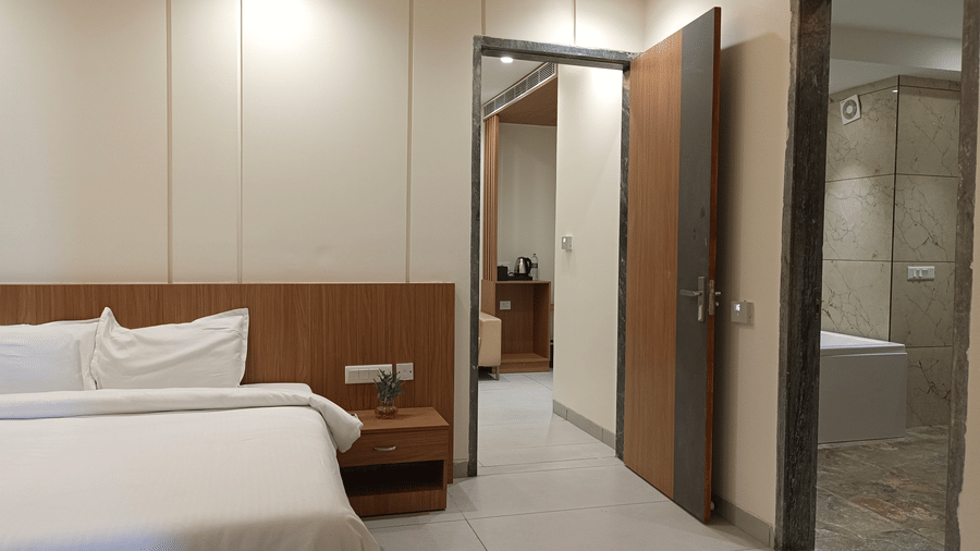 A view of the bedroom area of the Presidential Suite at The Prince ZENQ Xpress Resortt Rohtak, showing a large bed, wooden nightstand, and two open doorways leading to the suite's other areas, one of which shows a bathroom with a tub.