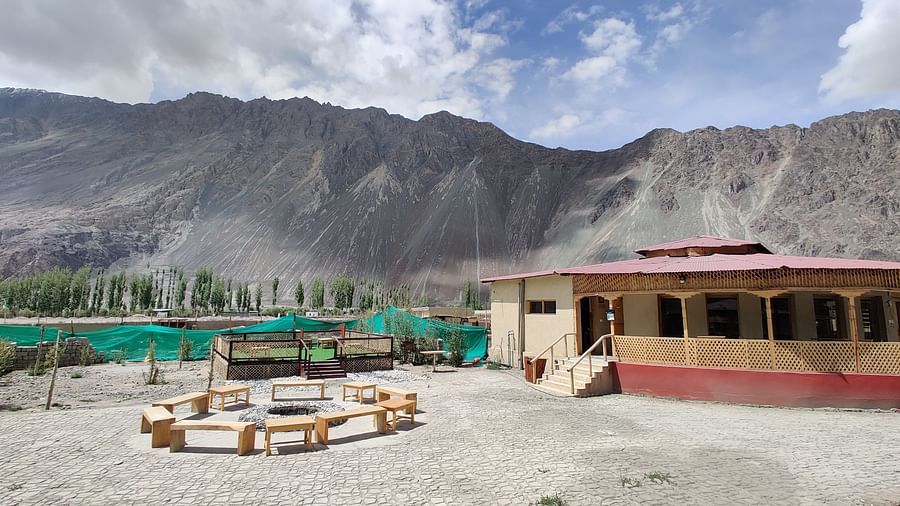 A rustic outdoor area with simple seating, set against a backdrop of rugged mountains and a cloudy sky at White Sands Resort, Ladakh.