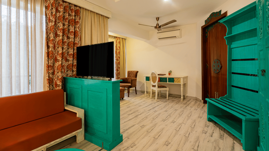 A room featuring a multi seater sofa, a television unit, an armchair, a spacious writing desk and a chair, a wooden door, and sheer and opaque curtains | Summit by the Ganges Beach Resort & Spa, Rishikesh