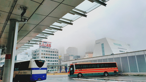 Two busses parked in a bus depot with buildings in the background