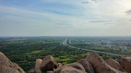 A panoramic view of a wide valley, with a highway visible in the distance, seen from a high point behind a rocky outcrop