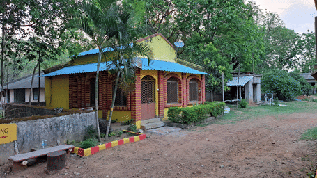 A yellow-fronted Family Cottage at Khairi Resorts standing next to a dirt path, with various trees and greenery surrounding it.