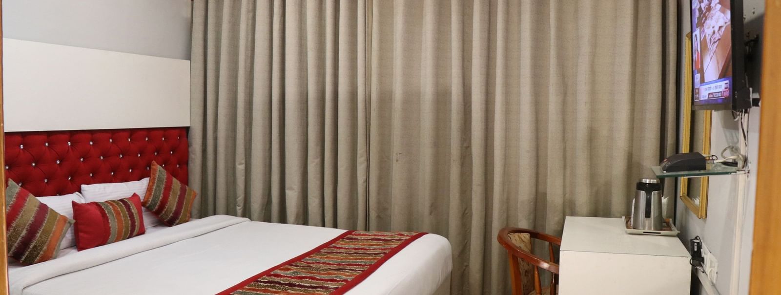 A hotel room with a plush bed, layered curtains with a red valance, a TV, a wooden chair, and an AC unit, illuminated by soft lighting. - Hotel Diamond Plaza, Chandigarh.