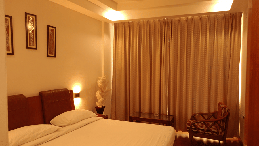 Guest room at Hotel Gold, Panipat featuring a bed, chair, side tables, and large window curtains.