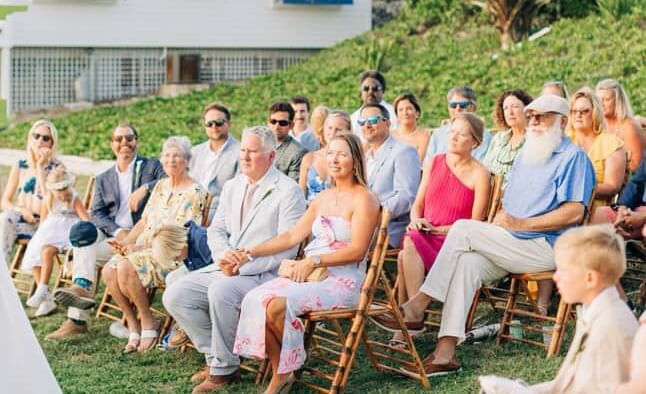 Wedding guests sit in rows of chairs on grass outside blue and white buildings at Abaco Inn.