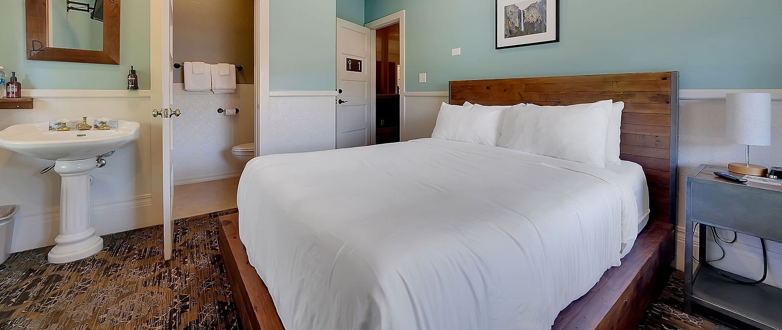 The Groveland Hotel's compact room features sage green walls, a wooden bed with crisp white bedding, and a pedestal sink with mirror.