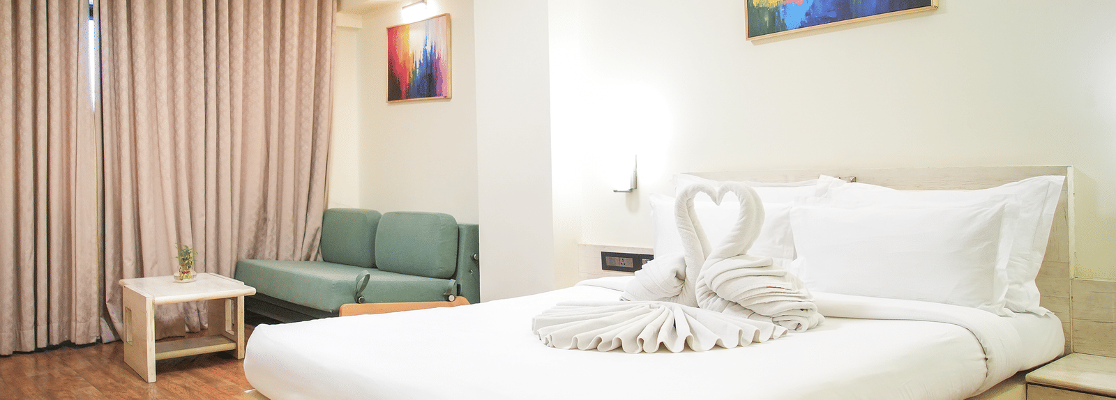 A room with a king size bed, a sofa and a chair, wall arts and light brown curtains at Infiniti Hotel, Indore.