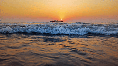A beach with a ship sailing in between waves, with the sky reflecting on the wet sand during sunset.