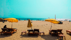 A sandy beach featuring several wooden lounge chairs arranged in rows under open yellow umbrellas, with distant figures visible near the ocean water line.