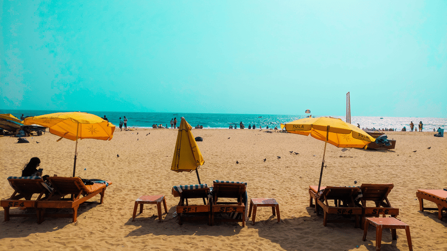 A sandy beach featuring several wooden lounge chairs arranged in rows under open yellow umbrellas, with distant figures visible near the ocean water line.