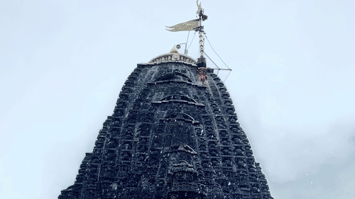 A tall stone temple tower viewed from below with a flag at the top against the sky with intricate art on the exterior.