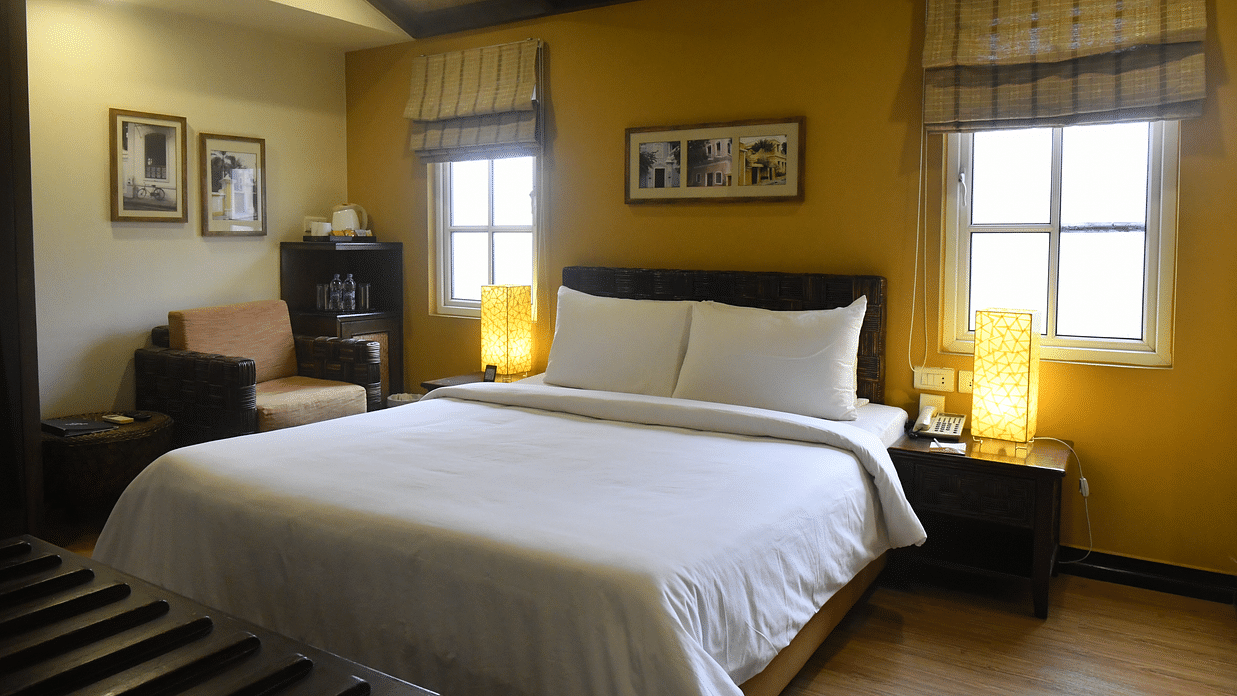 Deluxe Room at The Richmond, Puducherry featuring a plush bed with fresh linen and two lamps placed on the both sides on the nightstand as the sunlight makes its way through the two windows next to a sitting space
