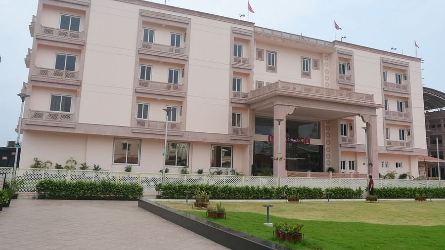 The front view of a modern, light-coloured hotel building with a covered entrance and green lawns in front is presented - Kankidham Lords Inn